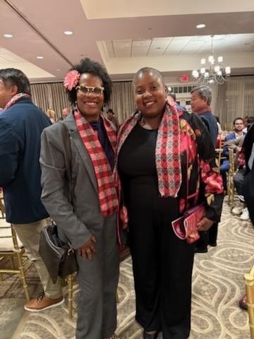 Two women smile for the camera at an indoor event. Both are wearing red and black patterned scarves, and one woman has a pink flower in her hair. The setting includes other attendees in the background, seated and standing in a warmly lit room with chandeliers.