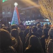 Large crowd gathered at night for a holiday celebration, facing a tall Christmas tree lit with red, green, blue, and white lights, as people look on during a festive tree-lighting event in a city square.