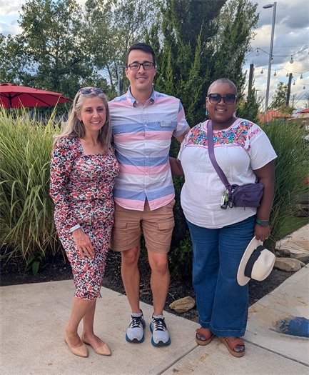 Three people stand smiling together outdoors in front of tall grasses and greenery. The person on the left wears a floral dress, the person in the middle wears a striped shirt and shorts, and the person on the right wears a colorful embroidered top, jeans, and holds a white hat.