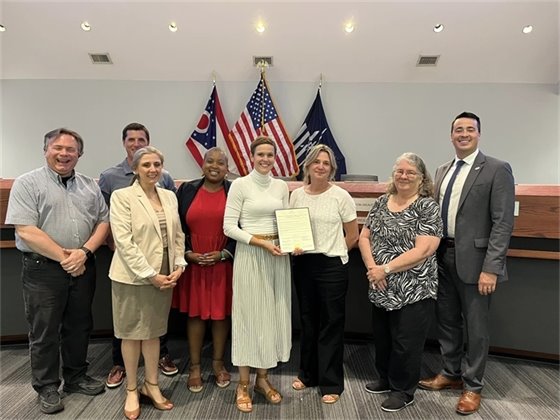 A group of eight people stand together in a council chamber, all smiling. One woman in the center holds a framed document. The backdrop features American and Ohio state flags, adding to the official setting of the photograph.