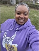 A smiling woman with short, salt-and-pepper hair takes a selfie outdoors in a grassy backyard with a wooden fence, a shed, and trees in the background. She is wearing a purple hoodie featuring a hand graphic with words that read "Include," "Love," "Listen," "Accept," and "Every Voice Counts" in a heart shape. She is also wearing silver hoop earrings.