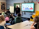 Councilmember Jamille Jones stands at the front of a classroom speaking to a group of elementary school students seated on the floor and at desks. A large screen behind her displays an educational video about the three branches of government: Executive (Governor), Legislative (Lawmakers), and Judicial (Judges). The classroom is decorated with educational posters, bookshelves, and a bright yellow reading chair. The students appear engaged and attentive.