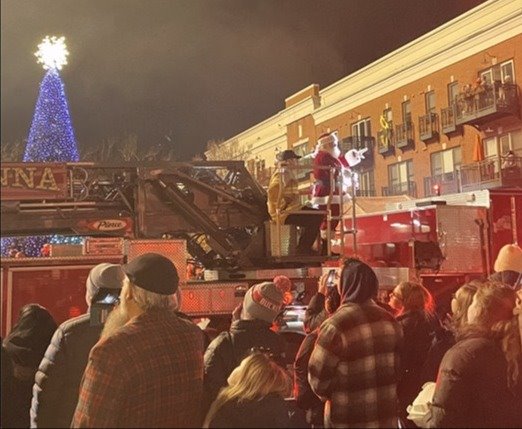 A festive crowd gathers outdoors at night, watching as Santa Claus waves from a Gahanna fire truck ladder. A brightly lit Christmas tree glows in the background.