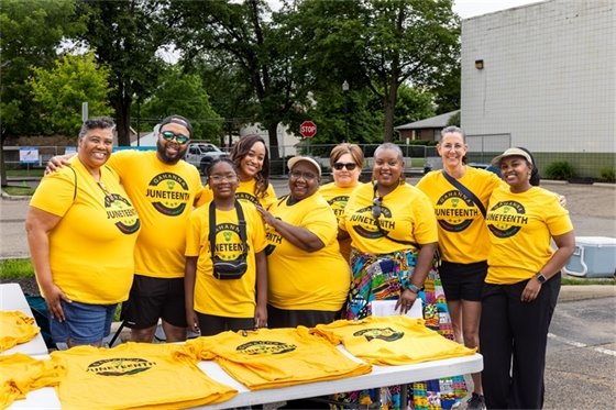 A group of people wearing matching yellow 'Gahanna Juneteenth' T-shirts smile and pose together behind a table covered with more event shirts. They are outdoors in a parking lot area, surrounded by trees and buildings, gathered in celebration of Gahanna’s first Juneteenth event.