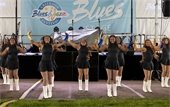 A group of young women in black outfits and white boots perform a choreographed dance on an outdoor stage. They wear sparkly gold and blue arm sleeves and smile as they strike a formation in front of a banner reading 'Blues & Jazz Festival Stage.