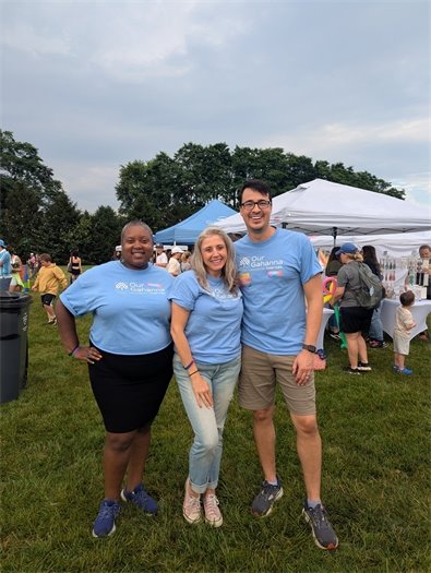 Three people wearing matching light blue event T-shirts pose together and smile at an outdoor community event. Behind them, families gather near white vendor tents on a grassy field under a partly cloudy sky.