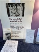  The image displays a poster titled "The Wonderful World of Herbs," promoting herb demonstrations on specific days and times at The Food Pantry Annex. The poster includes a black-and-white photograph of four women in vintage clothing holding herbs and related materials, likely from a historical exhibit by the Ohio Historical Society. Additionally, there are pamphlets and documents about herbs and their uses displayed on a table below the poster.