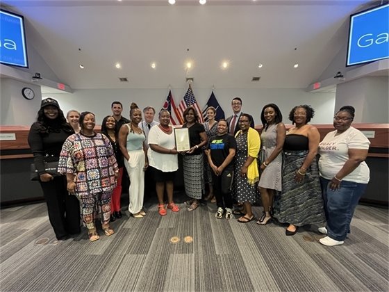 Group photo of councilmembers and community members in a meeting room, holding a framed certificate. Flags and monitors in the background display "Gahanna." Everyone is smiling and standing in a semicircle.