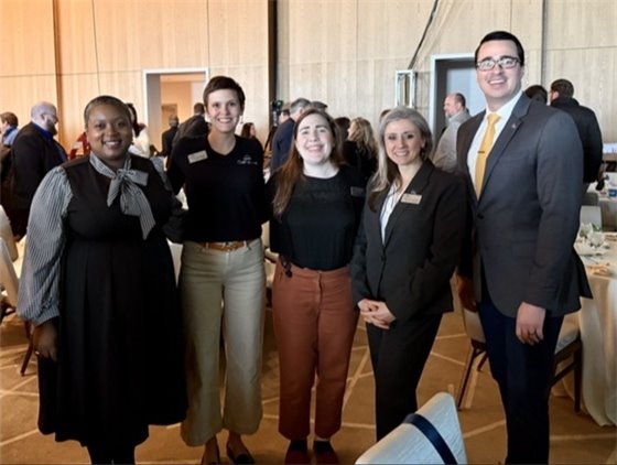 Five professionally dressed individuals stand together at a formal event with a wooden-paneled wall and other attendees mingling in the background. They are smiling and posing for the photo, with some wearing name tags indicating their roles or affiliations. The setting includes tables with white tablecloths, suggesting a conference or networking event.