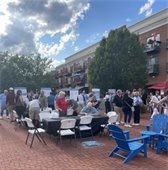 A large group of people gathers outdoors on a brick plaza with display boards set up around the space. Some attendees stand and read the boards while others sit at tables. Trees and a multi-story building line the background under a partly cloudy sky.
