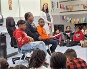 A classroom setting where a group of children sits cross-legged on the floor, attentively listening to a discussion. In the foreground, three individuals—two children and an adult—are seated in chairs, engaged in conversation. The adult, a woman wearing a dark-colored outfit with a name tag, is speaking, while the two children, one in a red hoodie and the other in an orange hoodie, are listening with papers in hand. A teacher or facilitator, dressed in a light-colored cardigan and a headband, stands in the background, smiling as she observes the interaction. The walls are decorated with student work, posters, and colorful hanging folders.