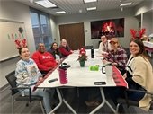 A group of seven people sitting around a festively decorated table in a holiday-themed room. Some are wearing holiday sweaters and reindeer antler headbands. A digital fireplace is displayed on a screen in the background, and small holiday decorations are visible on the table and walls.