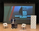 A man stands on stage in front of a large screen displaying the text "Real Men Real Talk Mental Wellness Summit 2024." The stage setup includes two white chairs, a small table, and a banner to the right. The man is wearing a navy jacket and sunglasses, addressing an audience visible in the foreground, some of whom are taking photos with their phones. The setting is an auditorium with a wooden floor and a red curtain backdrop, creating a formal yet engaging atmosphere for the event focused on mental wellness.