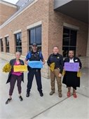 Four people standing in front of a brick building, each holding colorful signs with greetings like "Hey/Hola," "Hi! Heyyy," and "Waz up! Hola." Two of the individuals are police officers in uniform, while the other two are dressed in business casual attire and holding yellow pom-poms. They are all smiling.