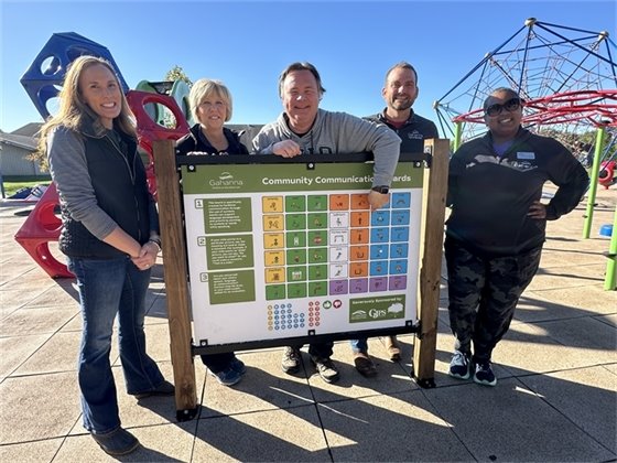 Photo from the ribbon-cutting event for new communication panels at Sunpoint Park. City council members, community leaders, and attendees celebrate the installation of panels designed to support nonspeaking and minimally speaking individuals in fully enjoying the park.