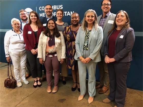 A group of eleven individuals standing together, smiling for a photo in front of a blue backdrop with the words "Columbus Metropolitan" partially visible. The group is diverse in gender, ethnicity, and attire, with a mix of formal and business-casual clothing. They appear to be at an indoor event or meeting, suggesting a professional or community gathering. Some individuals are wearing name tags.