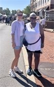 Two people smile while standing on a sunny street lined with vendor tents and a crowd at an outdoor market or community event. One wears a hat and sunglasses, and the other wears a striped shirt and shorts.