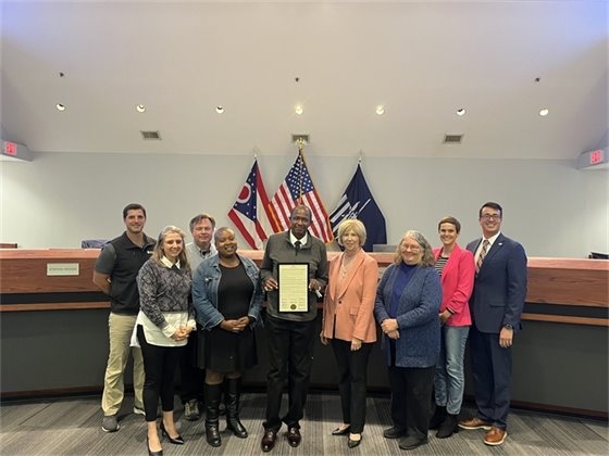 A group of people, including city council members and community leaders, standing together in a council chamber holding a framed proclamation, with flags of the United States and Ohio in the background.