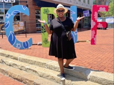  A woman wearing a black dress and hat poses in front of large, colorful "Gahanna" letters at Creekside Event Center. She smiles with arms raised, standing on brick steps.