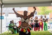 A woman dressed in colorful traditional African attire performs a joyful dance under a tent, arms raised and smiling. Behind her, other dancers and drummers in coordinated outfits watch and play instruments as part of a lively cultural performance at a Juneteenth celebration.