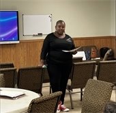 A woman stands in a room with rows of chairs, holding a notebook and speaking to a group. She is dressed in black with red sneakers and appears to be engaging with attendees, one of which is a child in the lower right corner of the image.