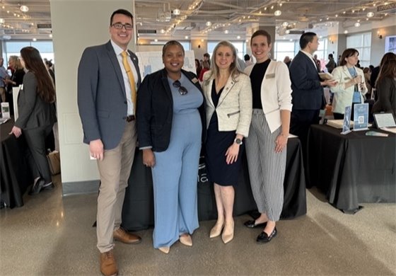 Four professionally dressed individuals pose together at an indoor event with large windows, string lights, and informational booths in the background. They are smiling and standing close together, with other attendees mingling and conversing behind them. The booths display materials related to Gahanna.