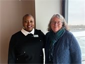 Two individuals standing side by side indoors, smiling warmly at the camera. The person on the left is wearing a black top with a white collar and a name badge, while the person on the right is wearing a blue sweater and scarf. A snowy landscape is visible through the window in the background.