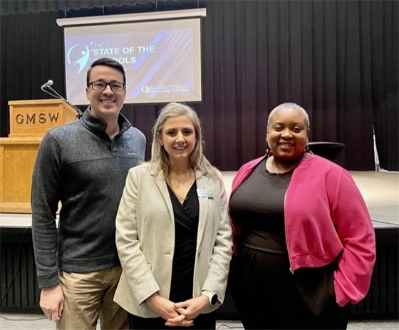 Three individuals standing together in front of a stage with a podium on the left and a screen displaying 'State of the Schools' in the background. The individuals are smiling, with the person on the right wearing a bright pink jacket, the person in the center wearing a light gray blazer, and the person on the left wearing a dark sweater.