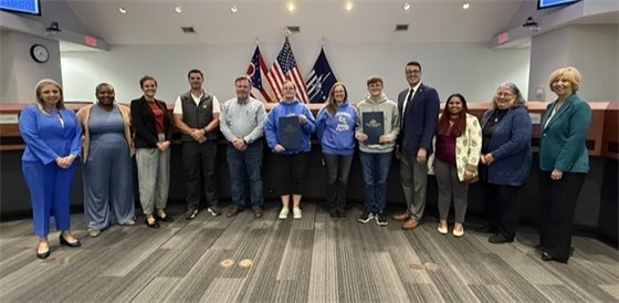 Two people smile while standing on a sunny street lined with vendor tents and a crowd at an outdoor market or community event. One wears a hat and sunglasses, and the other wears a striped shirt and shorts.