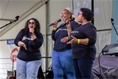 Three women wearing matching 'Harmony' shirts sing into microphones on stage, smiling and engaging the audience. They are performing under a tent at a community event, with music equipment and stage lights visible around them.
